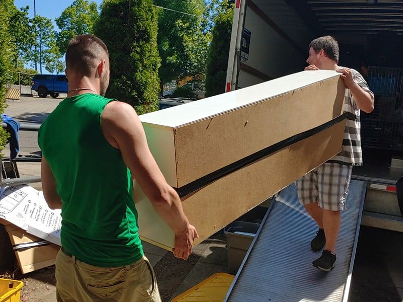 Two movers carrying a long boxed item down a truck ramp
